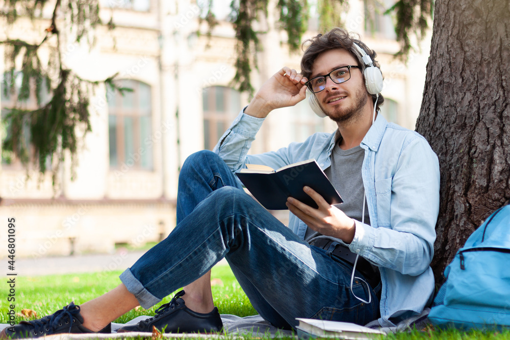 Happy student is sitting on green grass yard under tree, listening music, reading book outdoor at campus. Young smiling man is preparing for lessons, exam. Education concept. Back to high school.