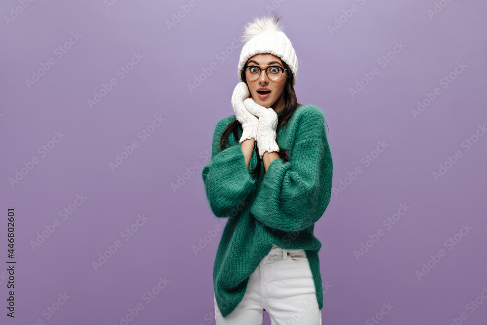 Surprised woman in green sweater and white pants poses on purple background. Young girl in knitted hat and gloves looks shocked on isolated. Surprised woman in green sweater and white pants poses on purple background. Young girl in knitted hat and gloves looks shocked on isolated.