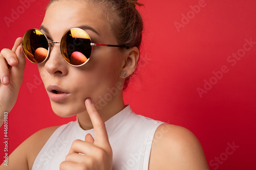 Charming positive surprised young blonde woman isolated over red background wall wearing casual white top and stylish colourful sunglasses looking to the side