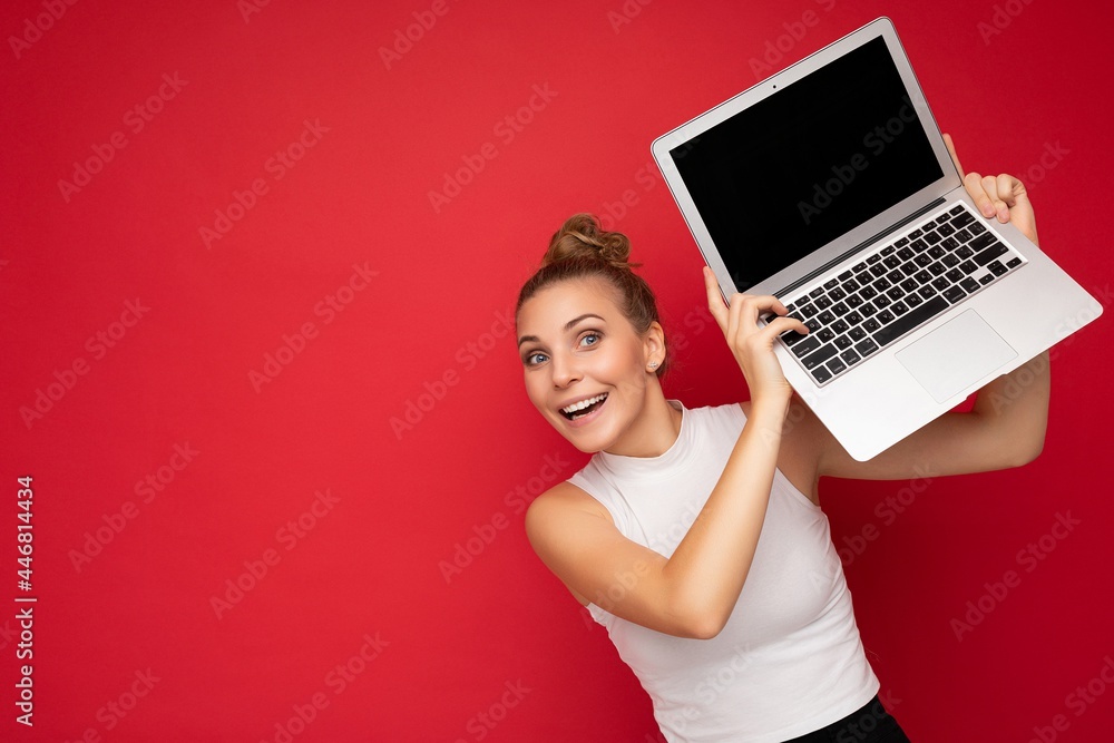 Naklejka premium beautiful fascinating happy blond young woman with open mouth with gathered hair looking at camera holding computer laptop wearing white t-shirt isolated over red wall background