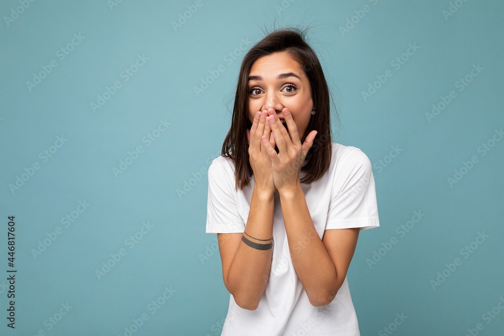 Portrait of positive surprised young beautiful brunette woman with sincere emotions wearing casual white t-shirt for mockup isolated on blue background with copy space and covering mouth with hands Portrait of positive surprised young beautiful brunette woman with sincere emotions wearing casual white t-shirt for mockup isolated on blue background with copy space and covering mouth with hands