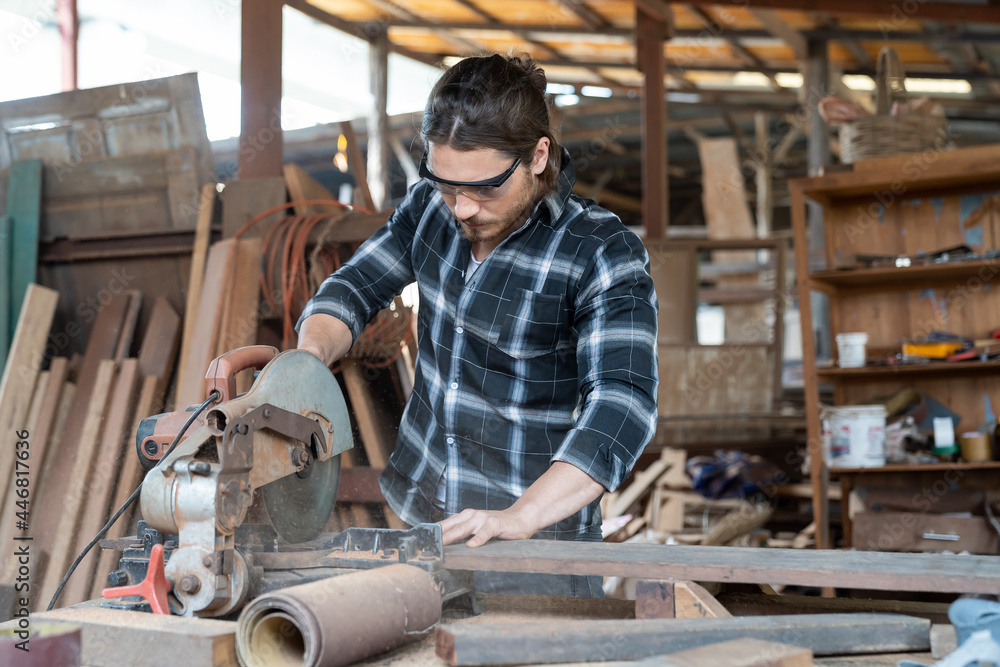 Male carpenter using circular saw cutting piece of wood and wearing safety goggles at woodwork workshop. Male woodworker working at work. Professional male craftsman Male carpenter using circular saw cutting piece of wood and wearing safety goggles at woodwork workshop. Male woodworker working at work. Professional male craftsman