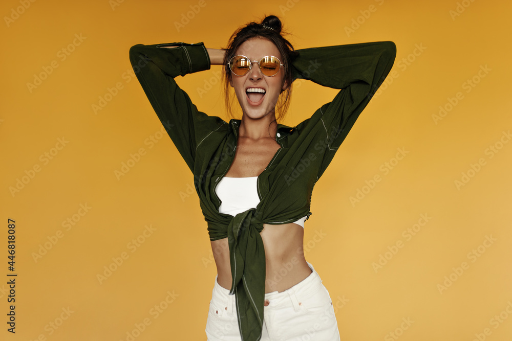 Cheerful woman in cropped top, shirt and white pants happily shouts on isolated. Portrait of emotional brunette girl in sunglasses on yellow background. Cheerful woman in cropped top, shirt and white pants happily shouts on isolated. Portrait of emotional brunette girl in sunglasses on yellow background.