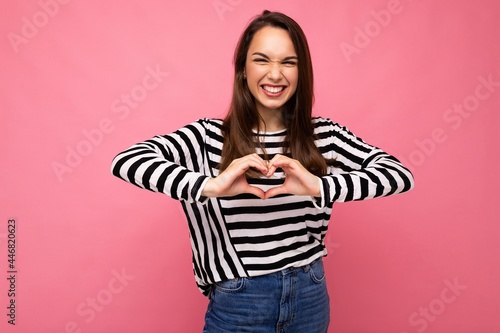 Young positive adorable happy amiling attractive brunette woman with sincere emotions wearing casual striped pullover isolated on pink background with copy space and showing heart gesture. Love photo