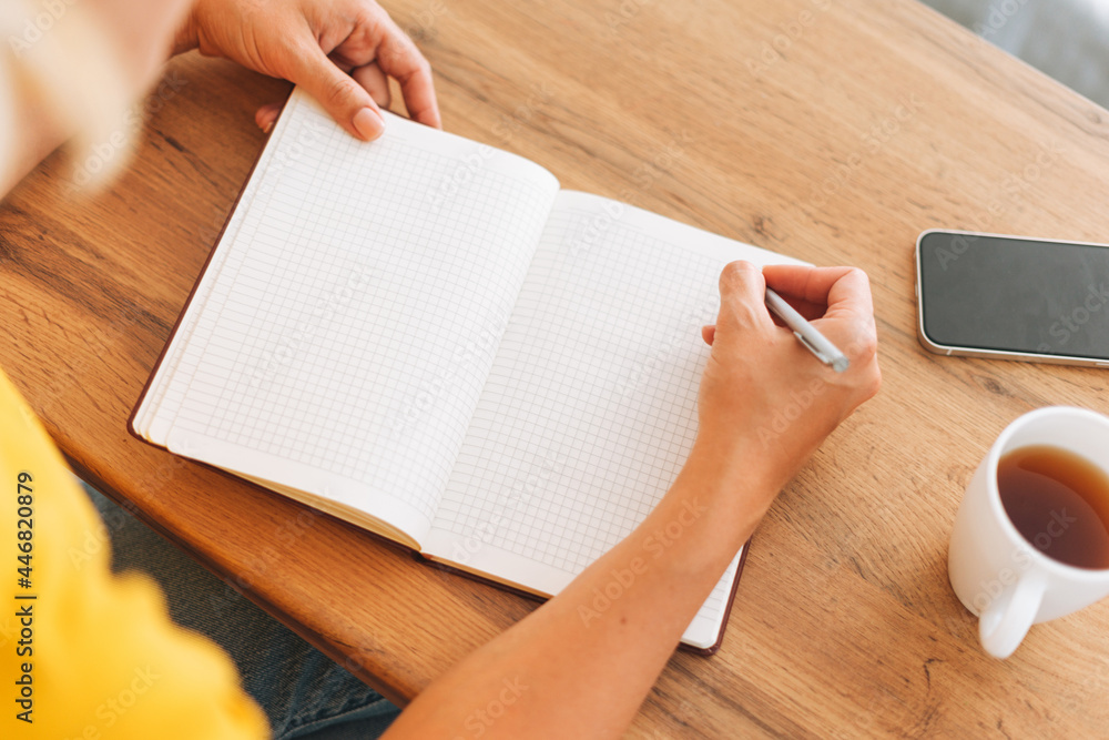 Young blonde woman in yellow t-shirt counts family budget and makes notes in notebook with cup of tea in the kitchen at home, top view Young blonde woman in yellow t-shirt counts family budget and makes notes in notebook with cup of tea in the kitchen at home, top view