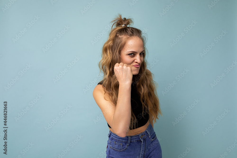 Young dissatisfied attractive winsome blonde curly woman with sincere emotions wearing trendy black top isolated over blue background with free space and showing fist. Negative concept