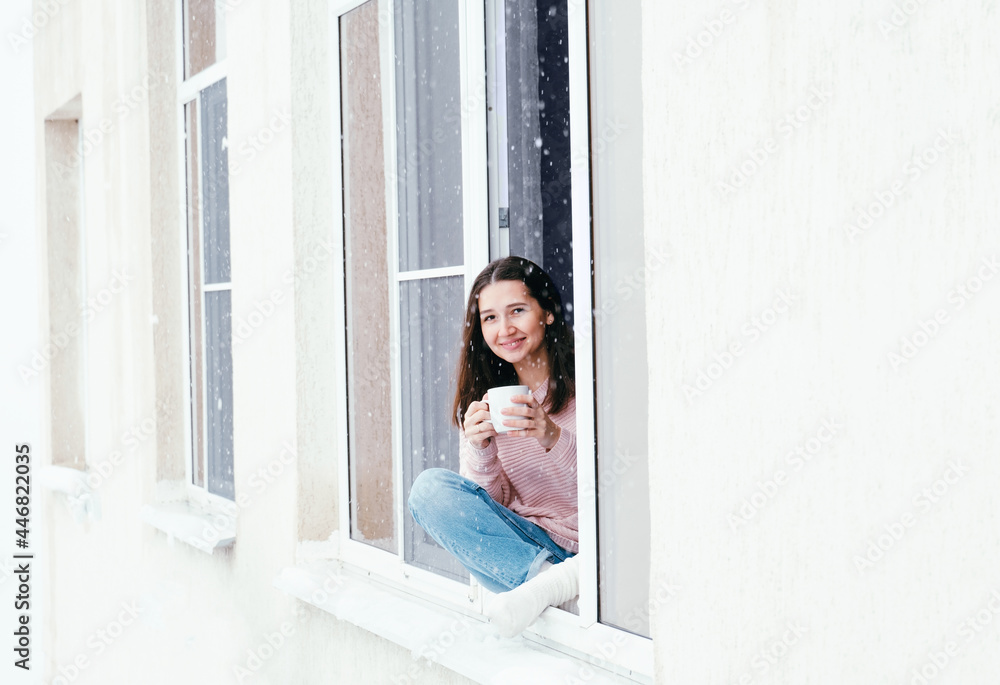 Beautiful brunette woman sitting on a window sill at home and drinking hot drink, cozy winter vacations portrait, copy space. Beautiful brunette woman sitting on a window sill at home and drinking hot drink, cozy winter vacations portrait, copy space.