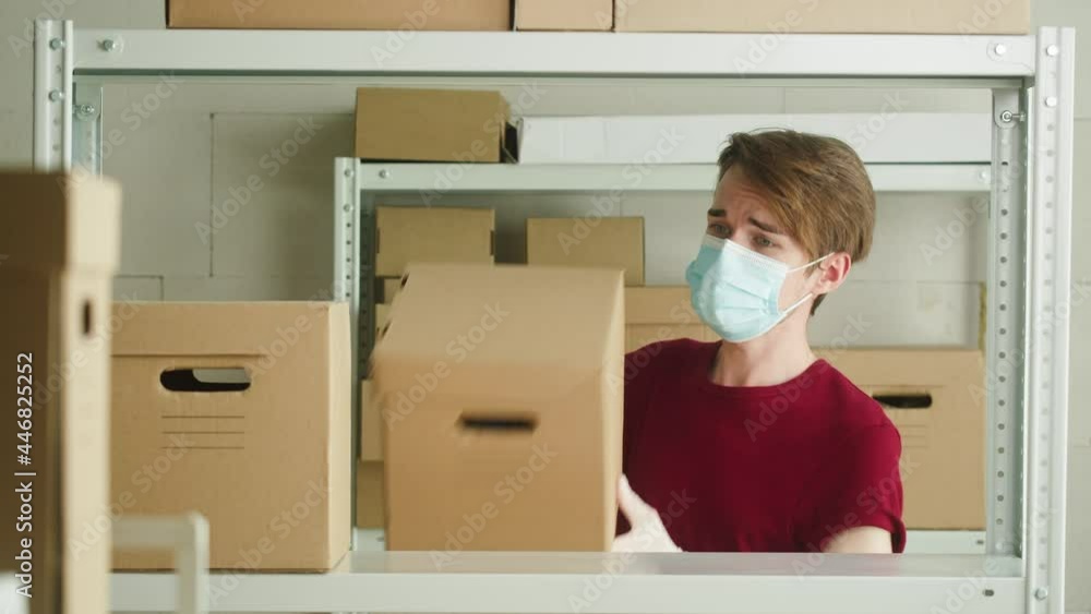 Man wearing medical face mask and protective gloves putting parcels on shelf, employee of warehouse puts delivery on the background of cardboard boxes in storage. Delivering during covid pandemic. Man wearing medical face mask and protective gloves putting parcels on shelf, employee of warehouse puts delivery on the background of cardboard boxes in storage. Delivering during covid pandemic.