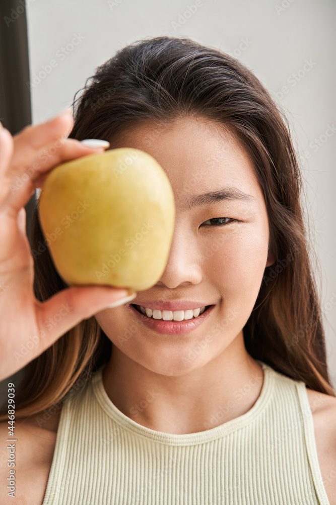Girl smiling toothy and looking at the camera while hiding her eye behind the apple Girl smiling toothy and looking at the camera while hiding her eye behind the apple