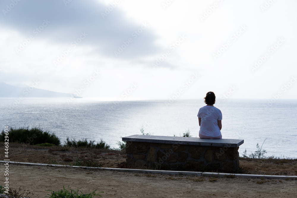 Woman sitting on a stone bench looking at the horizon on a sad and cloudy day