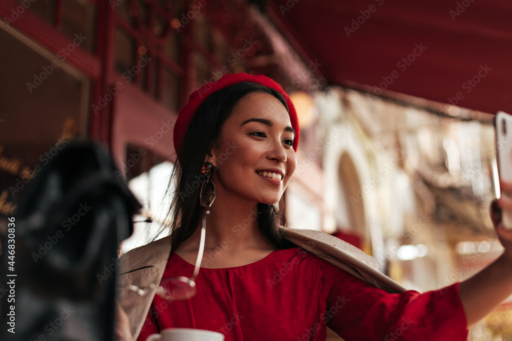 Happy young woman in red beret, stylish dress and beige trench coat holds eyeglasses, smiles and takes selfie in cozy street cafe.