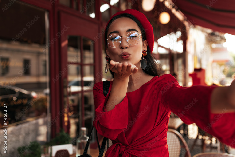 Fototapeta premium Happy Asian lady blows kiss outside. Cheerful brunette woman in beret and red dress takes selfie in street cafe.