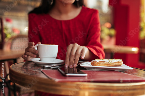 Photo of female tanned hands touching phone screen and holding white porcelain cup of coffee. Woman in red dress sits in cafe. Snapshot of tasty eclair on table.