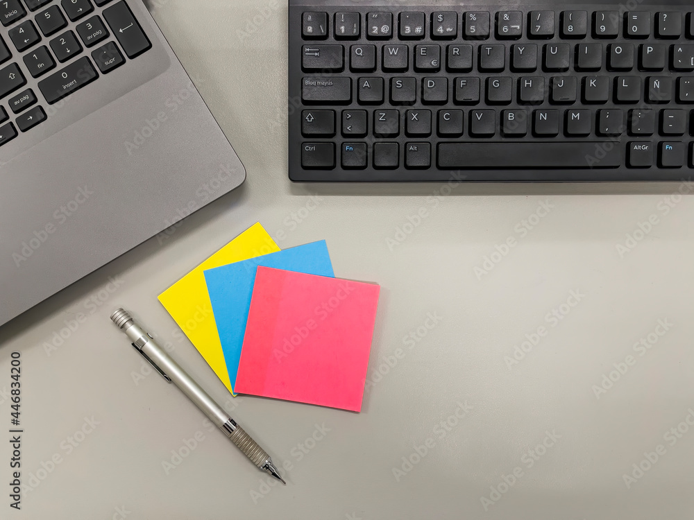 top view of a gray desk in an office with a black computer keyboard, a laptop keyboard, a mechanical pencil and three packets of yellow, blue and pink posit notes top view of a gray desk in an office with a black computer keyboard, a laptop keyboard, a mechanical pencil and three packets of yellow, blue and pink posit notes