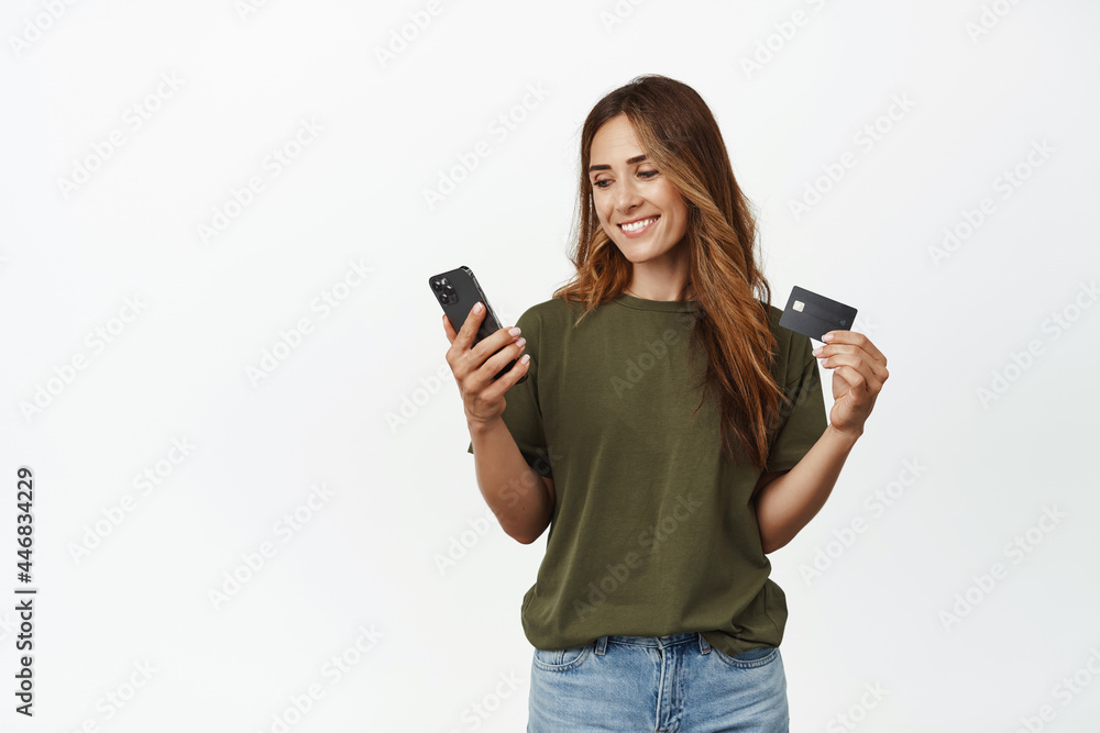 Smiling attractive middle aged woman, holding credit discount card, looking at smartphone screen, paying online, shopping in app, standing over white background Smiling attractive middle aged woman, holding credit discount card, looking at smartphone screen, paying online, shopping in app, standing over white background