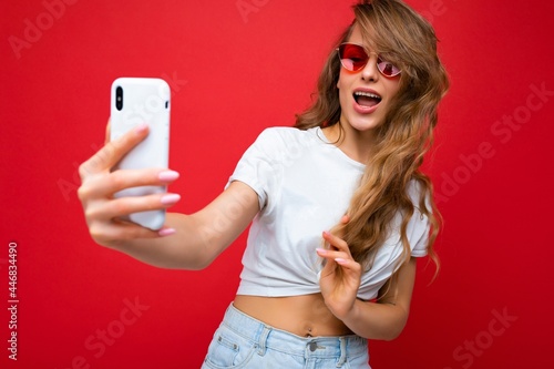 Closeup photo of sexy adult blonde female person holding mobile phone taking selfie photo using smartphone camera wearing sunglasses everyday stylish outfit isolated over colorful wall background