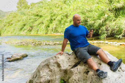 Chunky Caucasian hiker man using and making video calls on the phone sitting on a rock in a river. photo