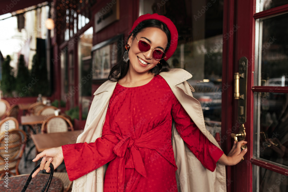 Pretty brunette elegant woman in red sunglasses smiles. Young lady in beret, bright dress and trench coat closes cafe door.