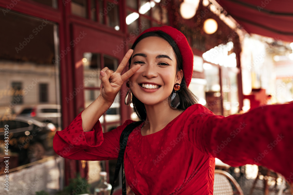 Happy charming brunette woman in red dress, stylish beret and eyeglasses smiles sincerely, shows peace sign and takes selfie outside. Happy charming brunette woman in red dress, stylish beret and eyeglasses smiles sincerely, shows peace sign and takes selfie outside.
