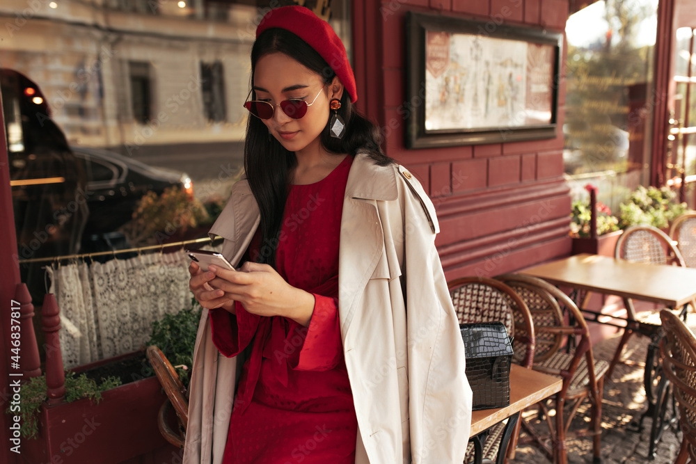 Pretty brunette woman in red dress, stylish sunglasses, beret and beige trench coat holds cellphone and leans on table in street cafe.