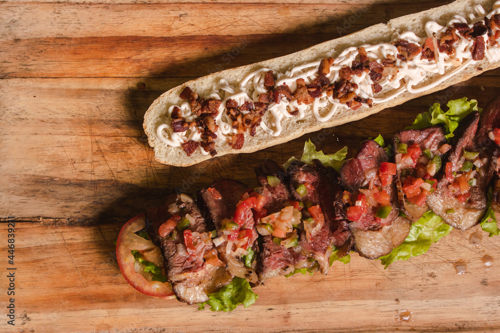 Take detail of a giant meat sandwich with vegetables on top of a wooden table with black background. Take detail of a giant meat sandwich with vegetables on top of a wooden table with black background.