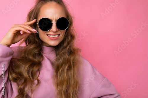 Closeup photo shot of beautiful smiling young dark blonde woman wearing casual clothes and stylish sunglasses isolated over colorful background looking at camera