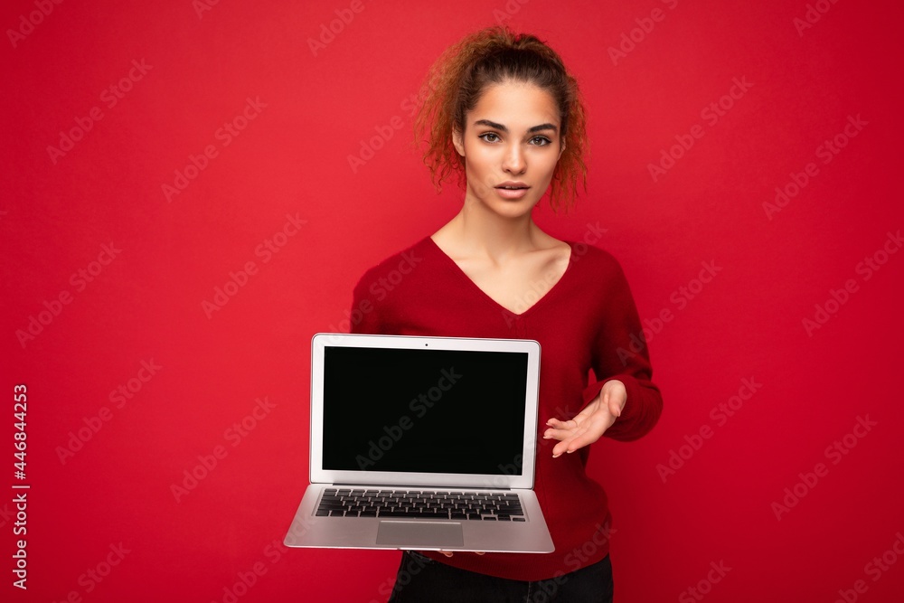Beautiful charming fascinating young curly dark blond woman wearing red sweater standing isolated over red wall background holding computer laptop with empty copy space mock up pointing to the netbook Beautiful charming fascinating young curly dark blond woman wearing red sweater standing isolated over red wall background holding computer laptop with empty copy space mock up pointing to the netbook