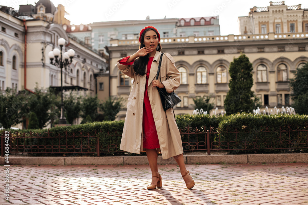 Young elegant lady in red dress, midi beige trench coat and beret poses outside and holds black leathered handbag.
