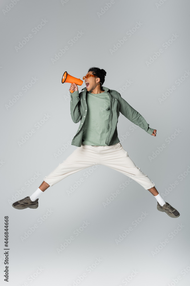 african american man in trendy clothes jumping with megaphone on grey background
