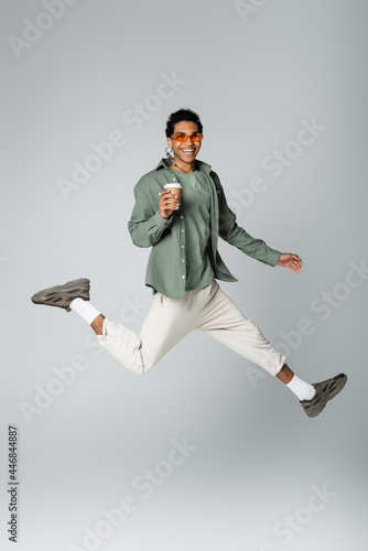 cheerful african american man in stylish clothes levitating with paper cup isolated on grey