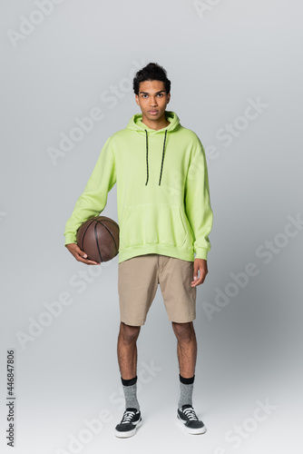 full length view of african american man in beige hoodie and green shorts holding basketball on grey background © LIGHTFIELD STUDIOS