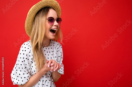 Photo shot of attractive happy smiling young blonde woman wearing summer casual clothes and stylish sunglasses isolated over colorful background wall looking to the side and having fun photo