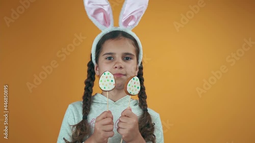 Happy Easter. Beautiful baby girl eats egg on a stick on a yellow background. Kid with rabbit ears eating Easter cookies.
