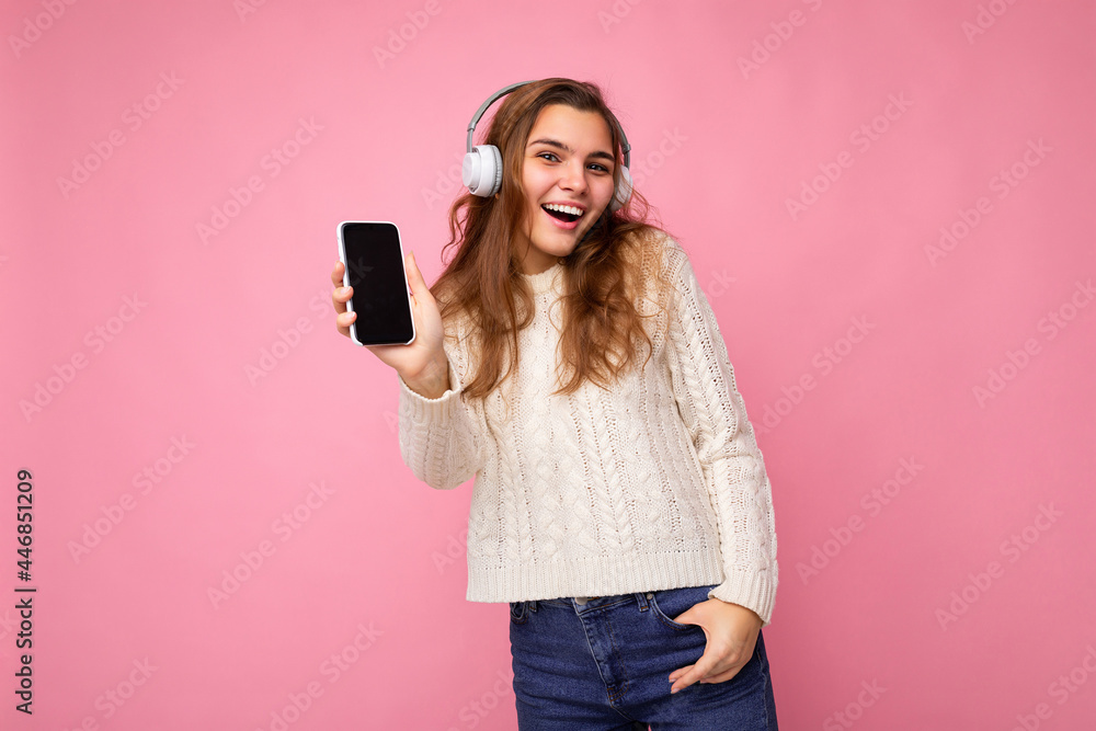 Fototapeta premium Beautiful happy smiling young woman wearing stylish casual outfit isolated on background wall holding and showing mobile phone with empty display for mockup wearing white bluetooth headphones