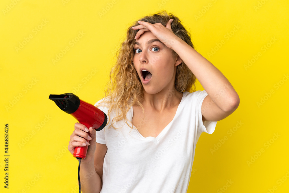 Young blonde woman holding a hairdryer isolated on yellow background doing surprise gesture while looking to the side Young blonde woman holding a hairdryer isolated on yellow background doing surprise gesture while looking to the side