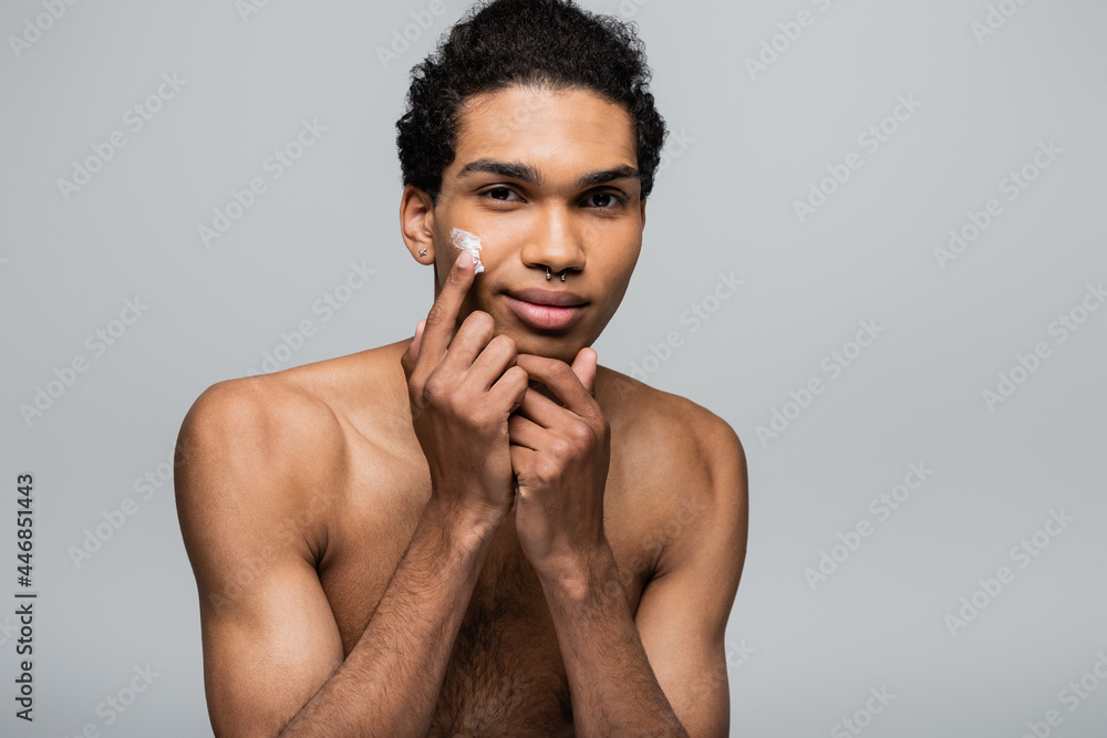 Fototapeta premium young african american man apply face cream while smiling at camera isolated on grey