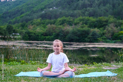 Beautiful happy girl with closed eyes practicing yoga in the lotus position near the lake in nature. Child learning fitness, stretching, yoga, active lifestyle concepts