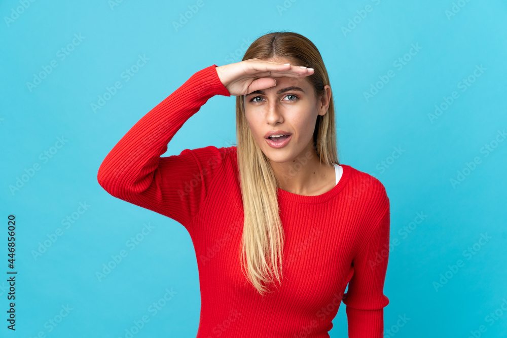 Young blonde woman isolated on blue background looking far away with hand to look something Young blonde woman isolated on blue background looking far away with hand to look something