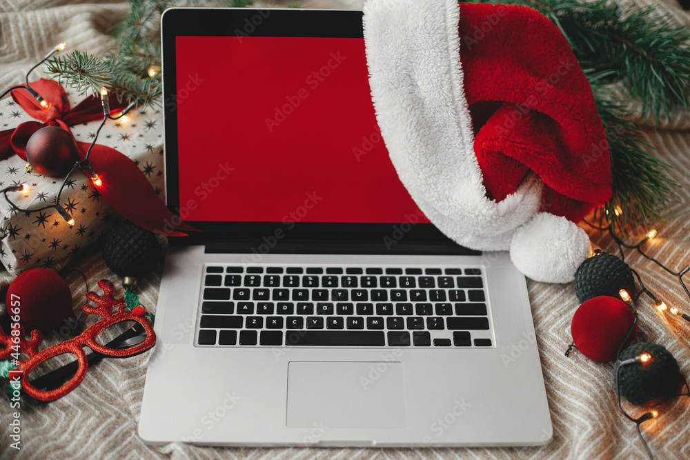 Christmas sales and shopping online. Laptop with red empty screen in santa hat on cozy bed with stylish christmas ornaments, present and festive lights. Space for text. View above. Freelance