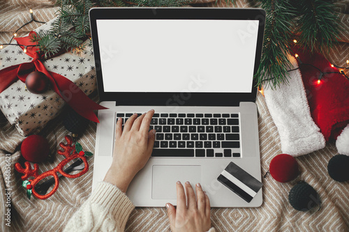 Christmas sales and shopping online. Hands on laptop with blank screen on cozy bed with credit card, stylish christmas ornaments, present, santa hat. Space for text. View above