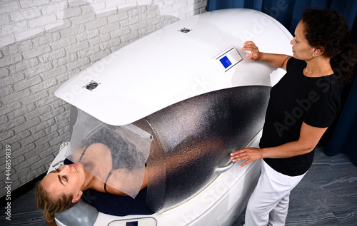 Doctor beautician adjusting programs in spa capsule with a woman inside it receiving professional beauty treatment
