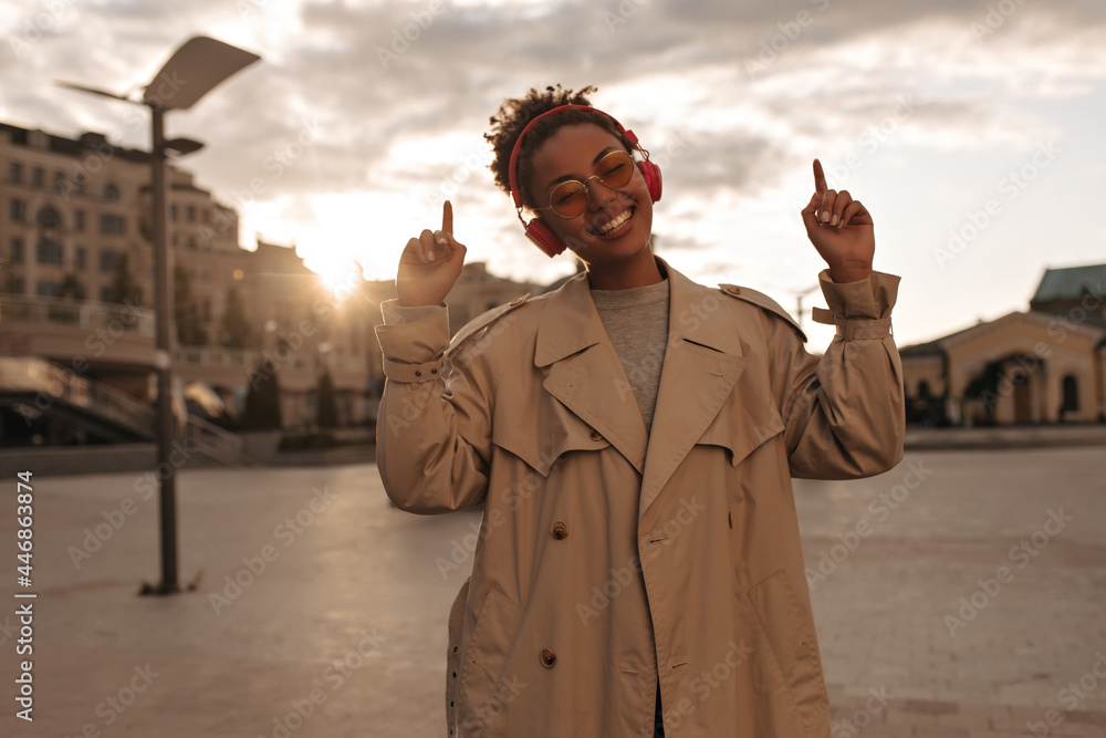 Curly dark-skinned lady in beige trench coat listens to music in headphones  outside. Charming woman in eyeglasses points up outdoors. Photos | Adobe  Stock