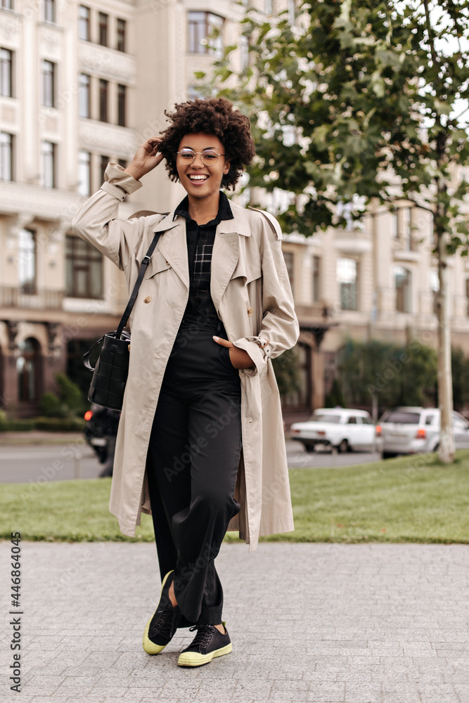 Joyful happy brunette curly dark-skinned woman in black pants, blouse and beige trench coat smiles and walks outside.