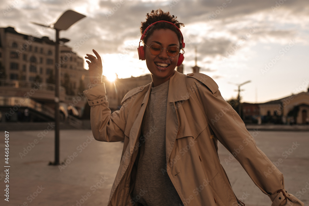 Stylish dark-skinned woman in beige trench coat dancing, smiling and listening to music in red headphones outside. Stylish dark-skinned woman in beige trench coat dancing, smiling and listening to music in red headphones outside.