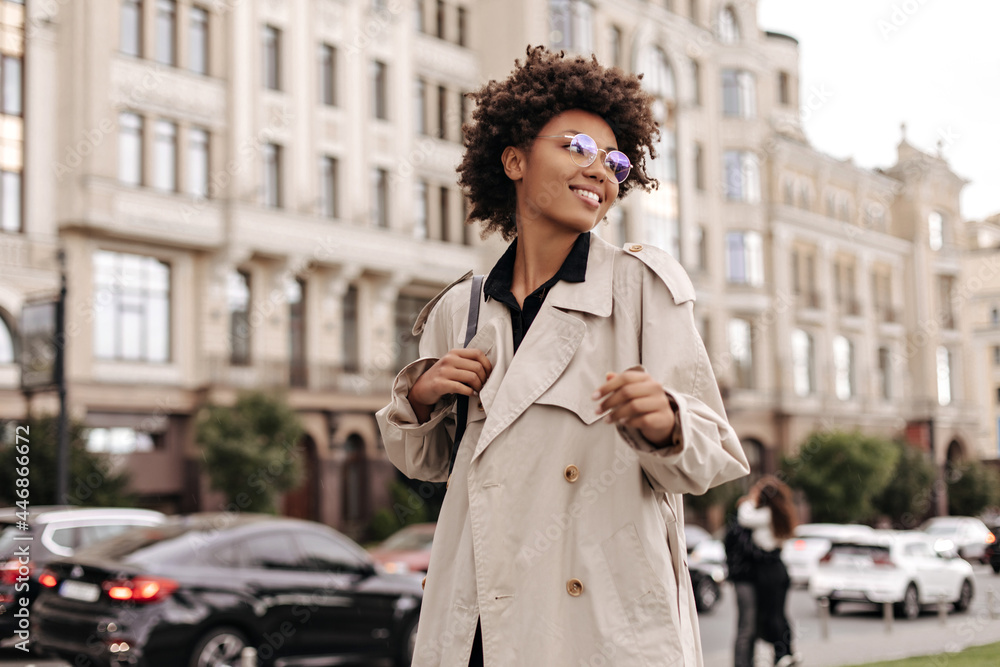 Fototapeta premium Excited brunette happy dark-skinned woman in eyeglasses, beige oversized trench coat smiles and walks outdoors.