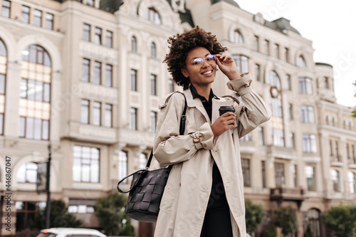 Wallpaper Mural Attractive cheerful dark-skinned woman in beige trench coat, black dress puts on eyeglasses, smiles, holds coffee cup outdoors. Torontodigital.ca