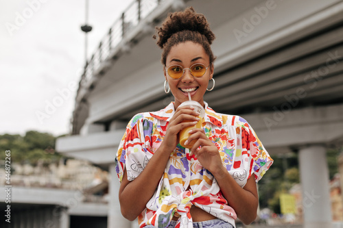 Excited curly brunette dark-skinned African woman in colorful blouse and orange sunglasses drinks cold juice and looks surprised outside.