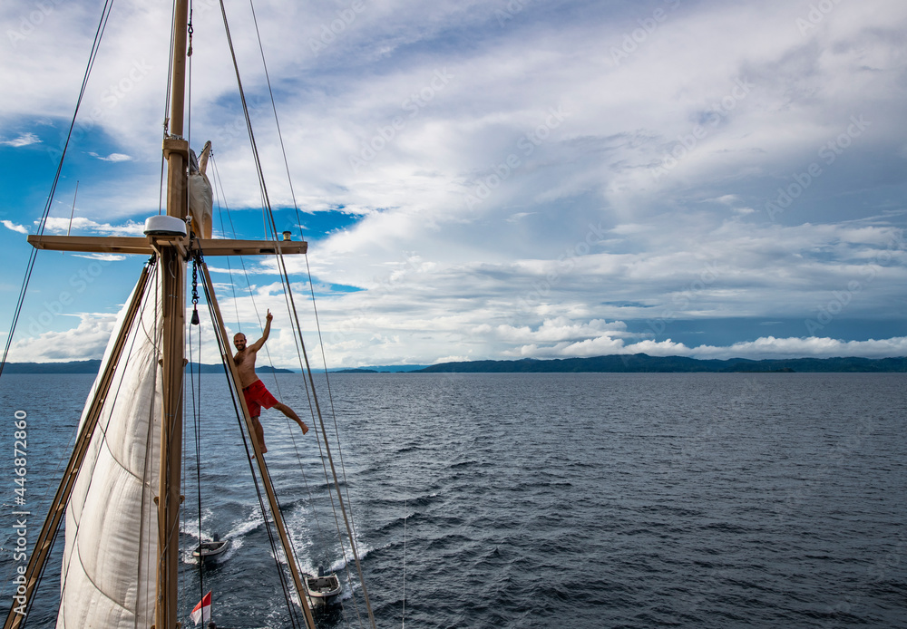 man in board shorts climbing up rigging of sail boat in Raja Ampat man in board shorts climbing up rigging of sail boat in Raja Ampat