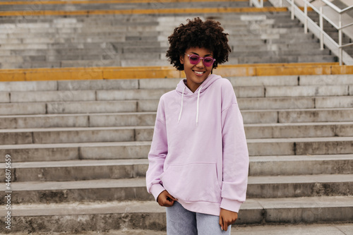 Charming pretty dark-skinned curly brunette woman in pink sunglasses and purple hoodie smiles and walks near stairs outside.