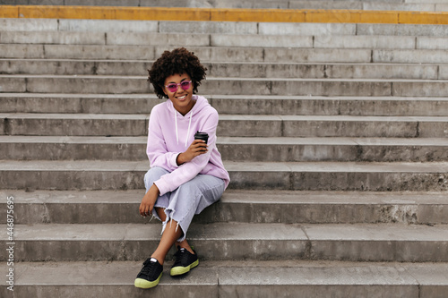 Joyful happy brunette curly dark-skinned woman in stylish jeans, purple hoodie and pink sunglasses smiles sincerely, holds coffee cup and sits on stairs outside. © Look!